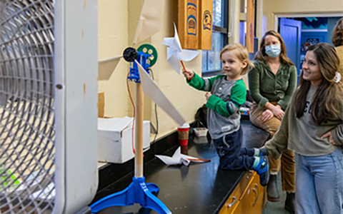 A child holds a paper windmill in front of a box fan.