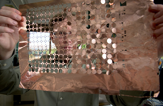 A person holds a sheet of copper discs in front of their face. Some of the sheet has empty holes where the discs have already come out.