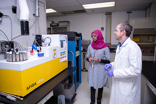 Two people in a fuels and combustion lab at NREL looking at a rectangular fuel analysis machine.