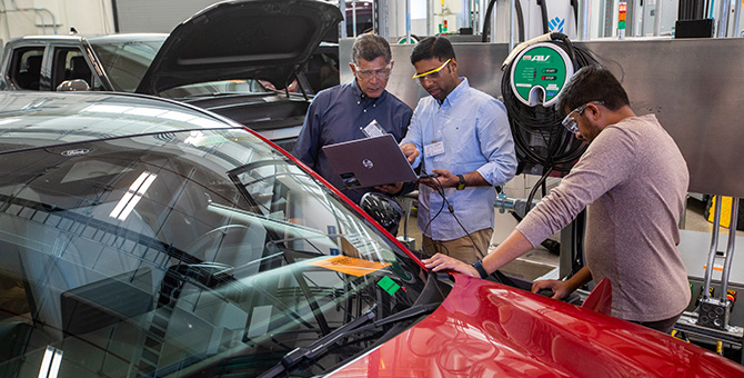 Three people standing around a red electric vehicle. One is holding a laptop and showing the screen to a second person, and a third person is plugging a charger into the vehicle.