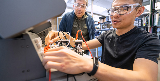 A person in the foreground handling wires and circuits while a person in the background looks on.