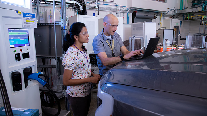 Two people look at a laptop sitting on top of the hood of a vehicle in front of an electric vehicle charger in an indoor laboratory.