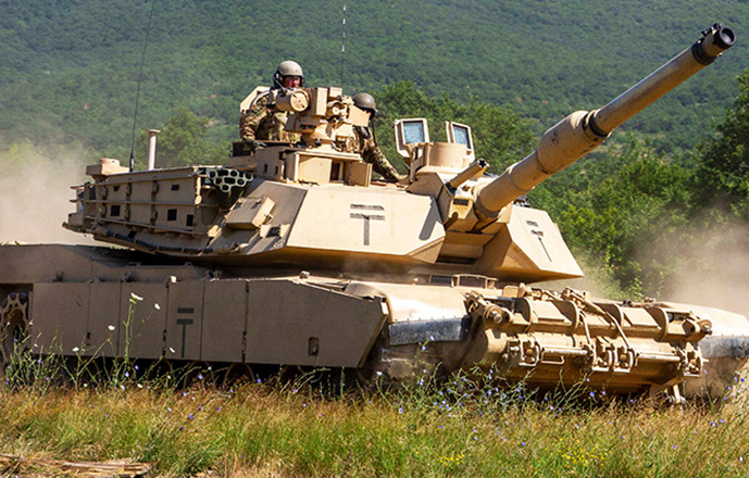 Several U.S. soldiers drive an M1 Abrams tank across a green field, kicking up a cloud of dust.