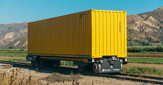 Photo of a yellow battery-electric, self-propelling rail car against a mountain background.