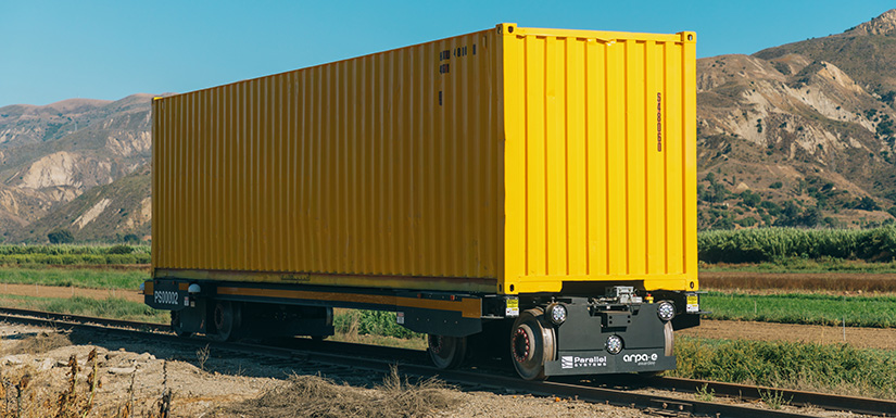 Photo of a yellow battery-electric, self-propelling rail car against a mountain background.