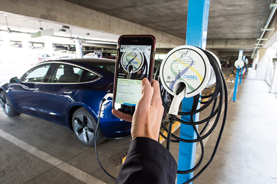 A person's hand holding a smartphone showing a smart charge management app in front of a plugged-in blue electric car.