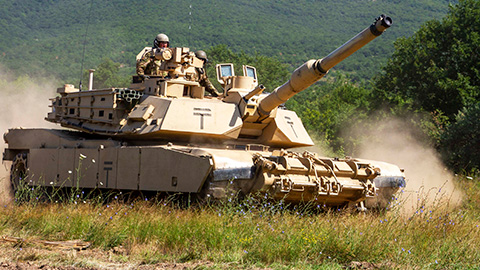 Several U.S. soldiers driving an M1 Abrams tank across a green field, kicking up a cloud of dust.