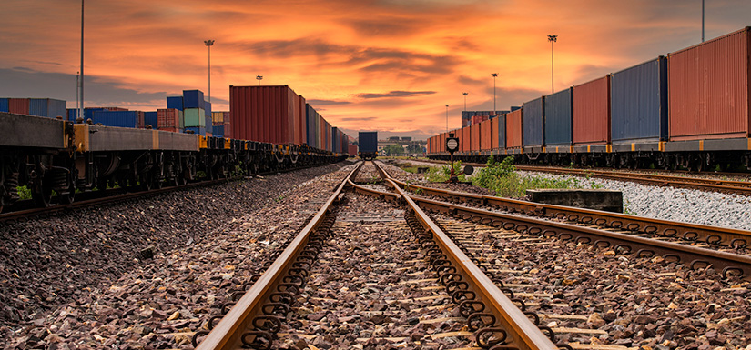 Two train tracks converge against a sunset. Cargo trains carrying intermodal shipping containers are visible on both sides.