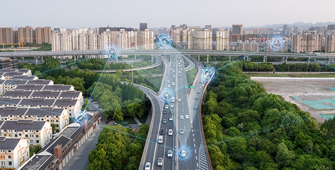 An aerial view of a highway flanked by trees and many nearby buildings. Some of the vehicles have blue circles and lines connecting them with each other, rooftop solar panels, and buildings.