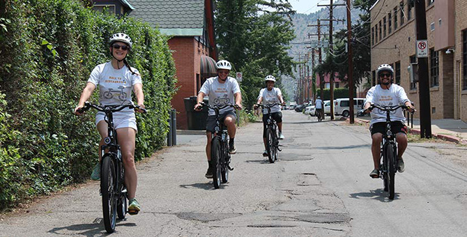 Four people wearing white shirts riding electric bicycles toward the camera and smiling on a narrow paved road flanked by greenery and buildings. 