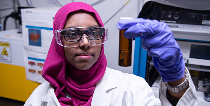  A person wearing a headscarf, safety glasses, gloves, and a lab coat holds up a test tube.