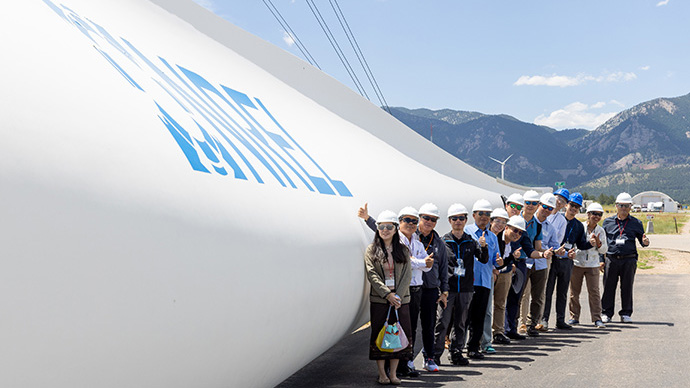 A group of individuals in hard hats smile and give a “thumbs up” as they stand in front of a large wind turbine blade with the NREL logo on it.