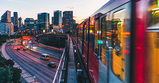 Train traveling above freeway in a city at dusk.