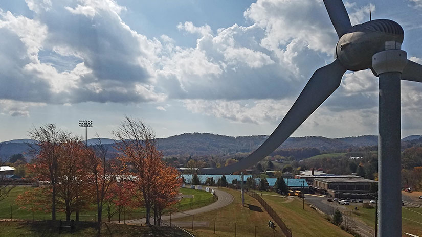 A small wind turbine against a background of fields, trees, a lake, and foothills