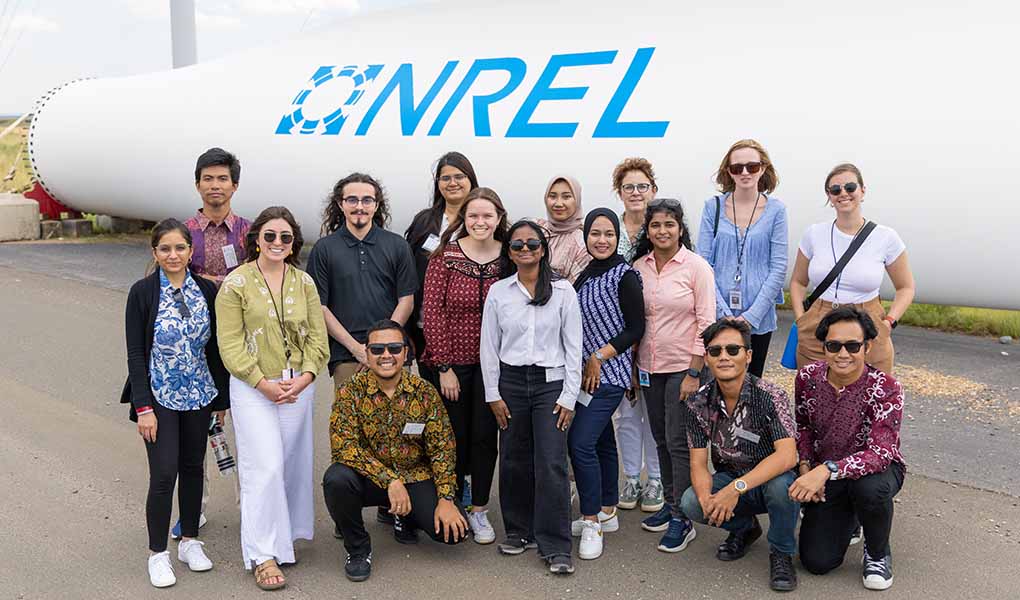 A group pose in front of a large wind turbine blade with the NREL logo on it.